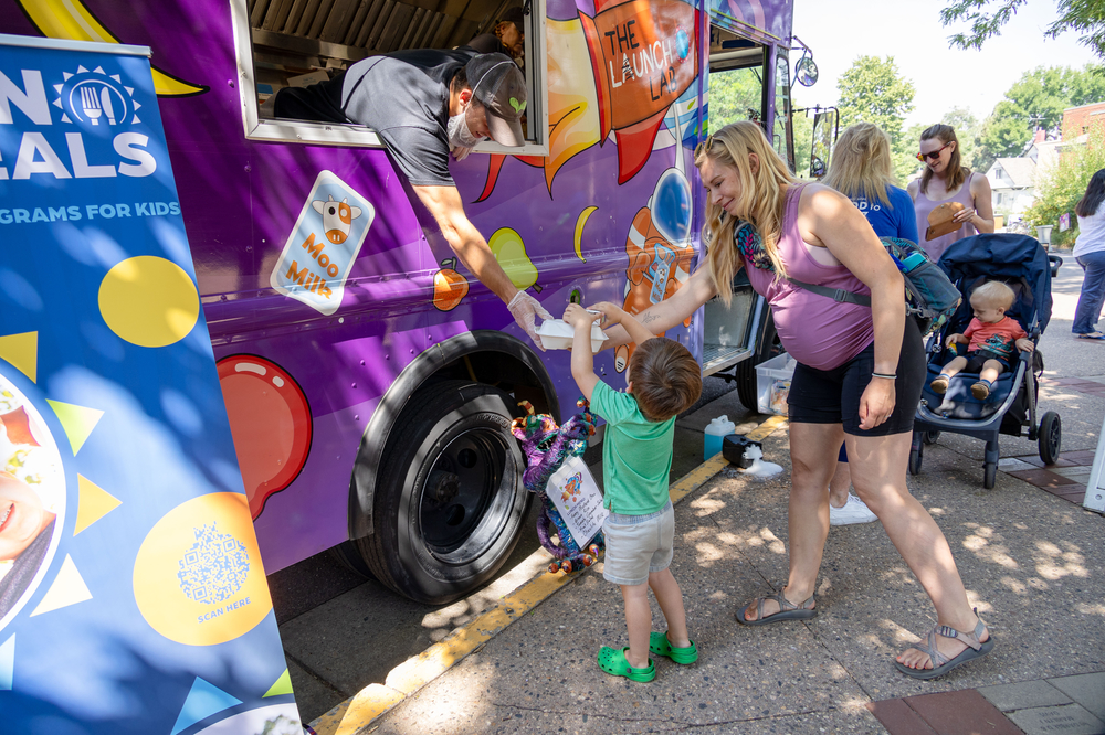 A small child with pregnant mother reaches up to a food truck for a box of food.